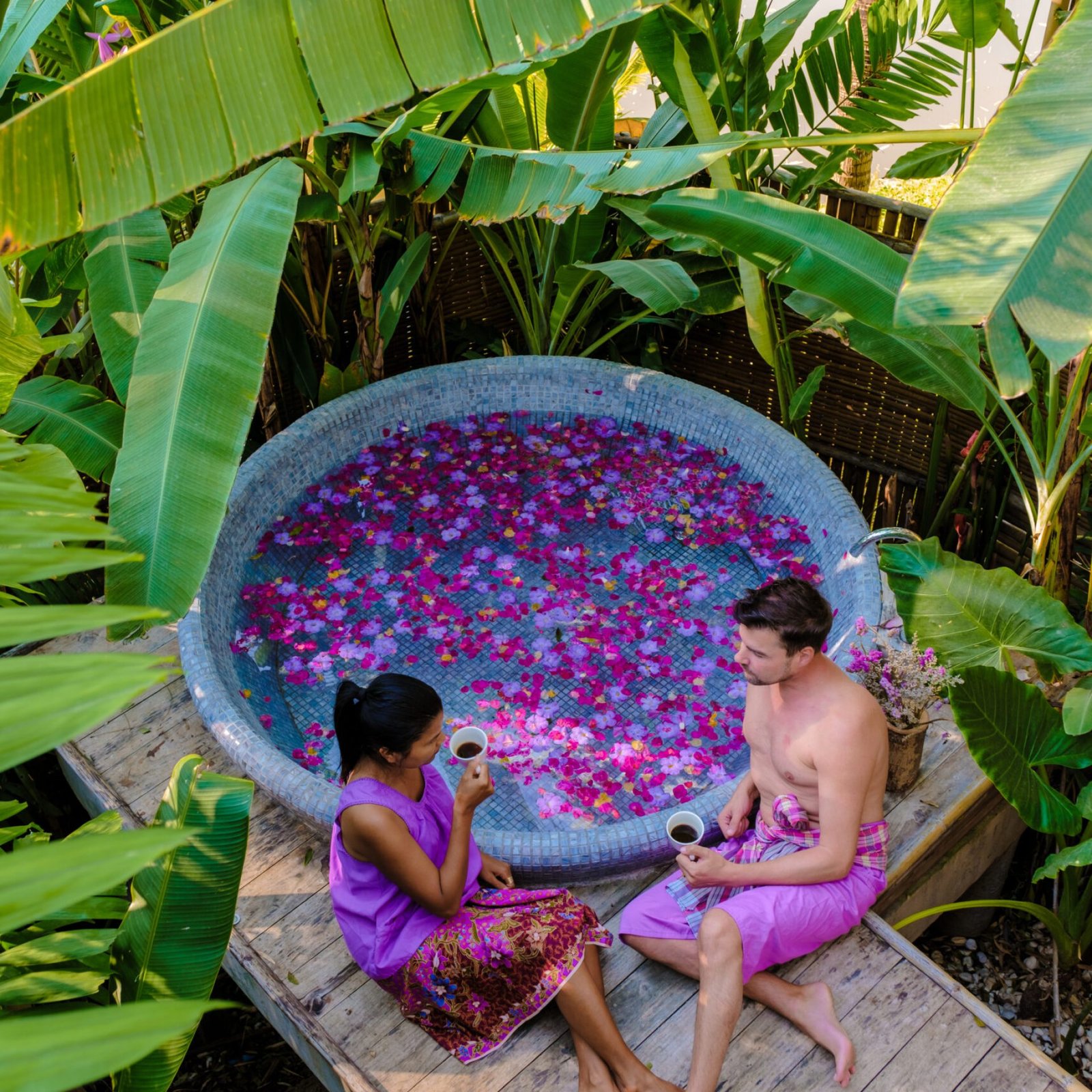 Couple at a bathtub in the rainforest of Thailand during a vacation with flowers in the bath, men and women relaxing in a jacuzzi with rose petals outside in nature and drinking coffee