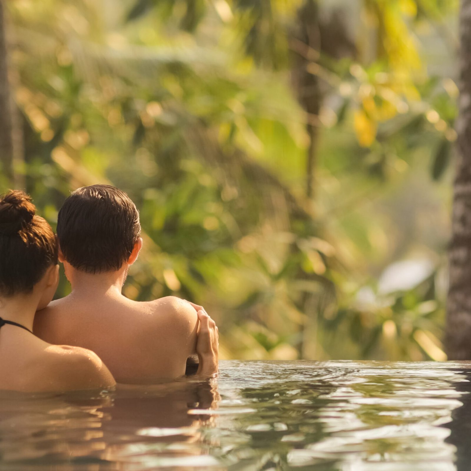 Panoramic photo of young adult couple in love swimming together in open-air infinity pool enjoying tropical vacation on villa looking away at rainforest on sunset. High quality photo